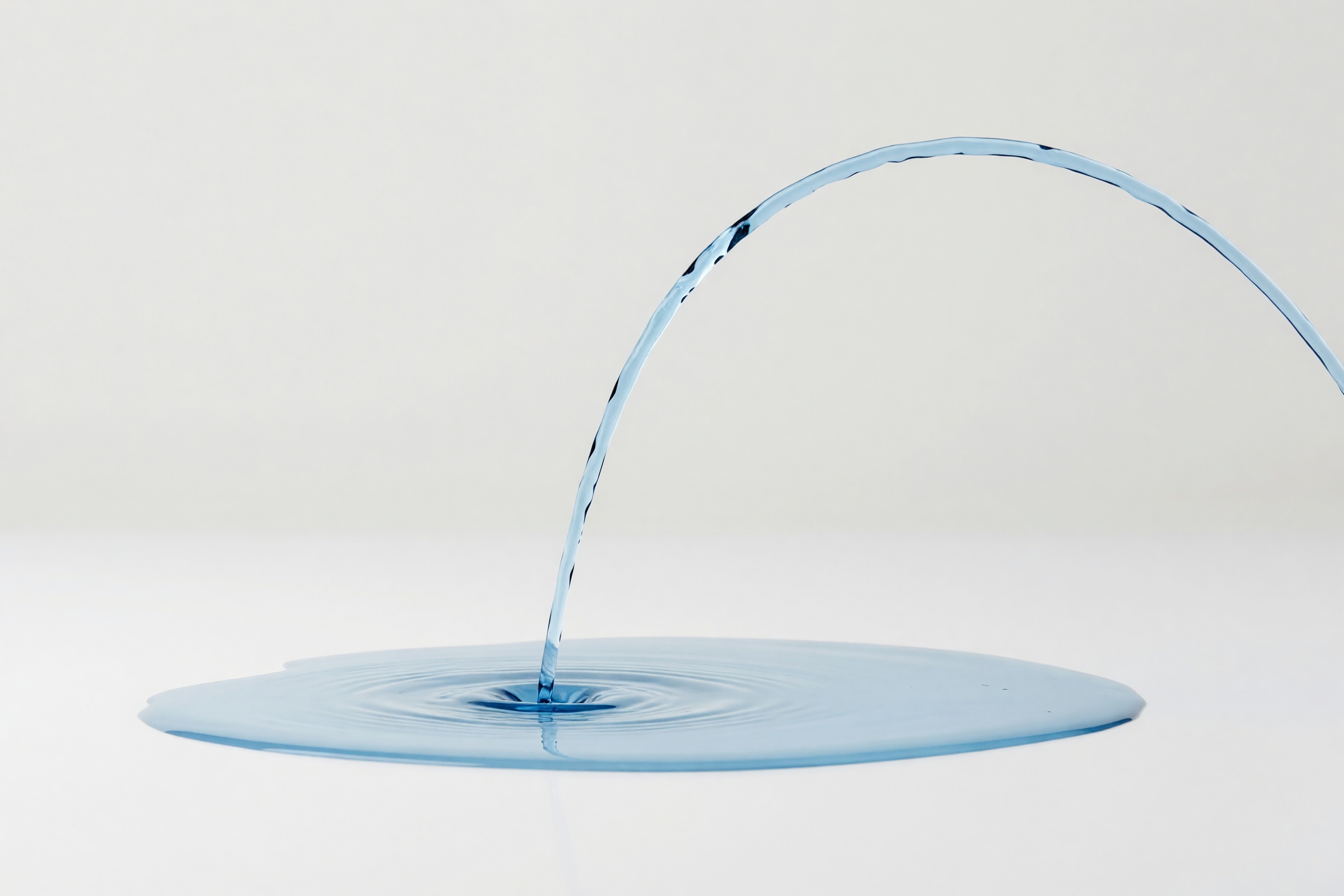 A smooth, thin arc of clear pale-blue liquid pouring into a shallow pool, on a clean studio ground