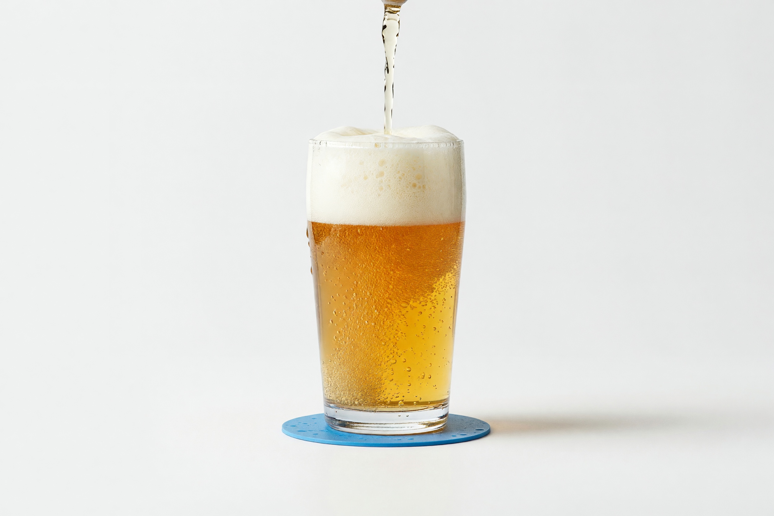 Golden beer with a frothy head pouring into a clear glass on a blue coaster, on a clean studio ground