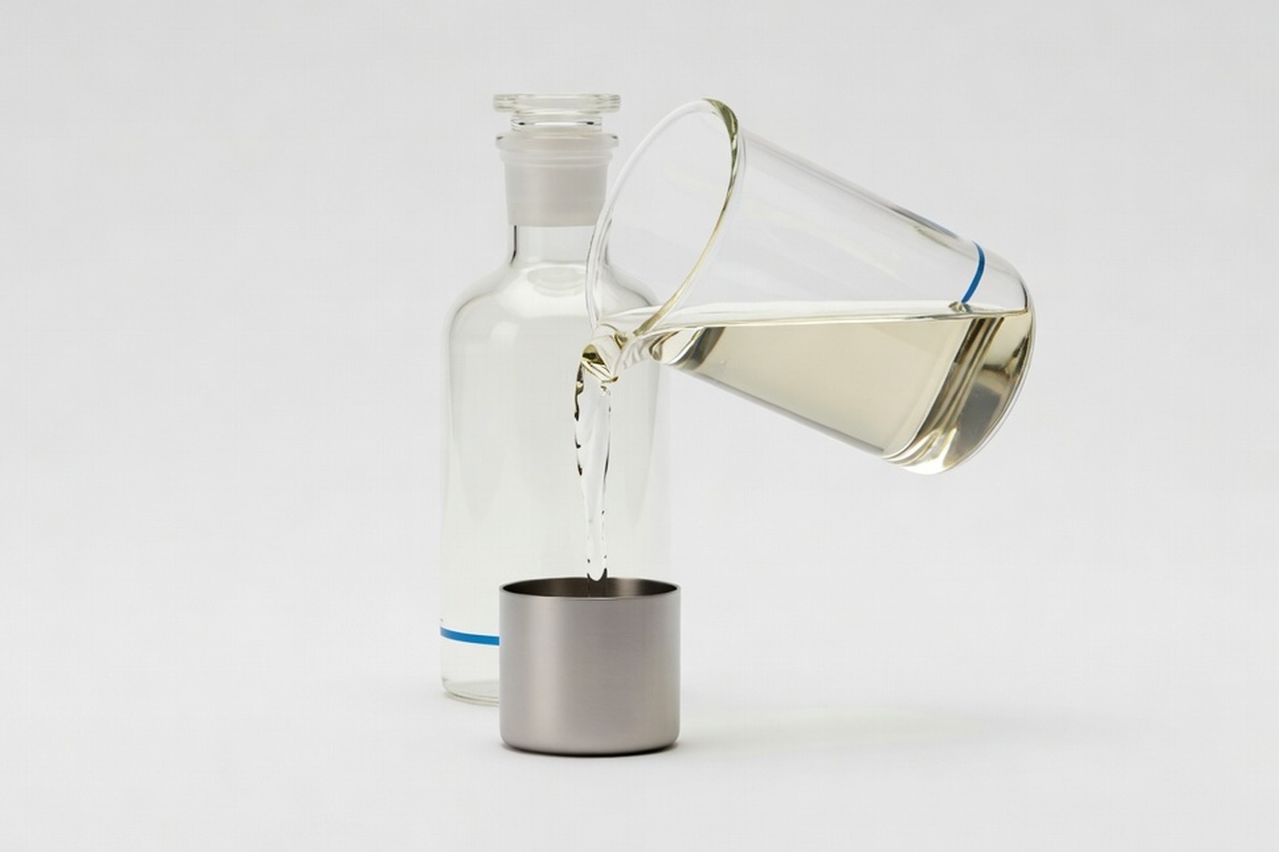 Clear chemical liquid pouring from a glass beaker into a stainless steel vessel on a near-white studio ground